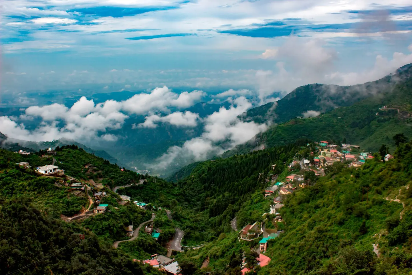 The Doon valley from a forested ridge, a hairpin road on the left, roofs down the slope and low cloud across the middle