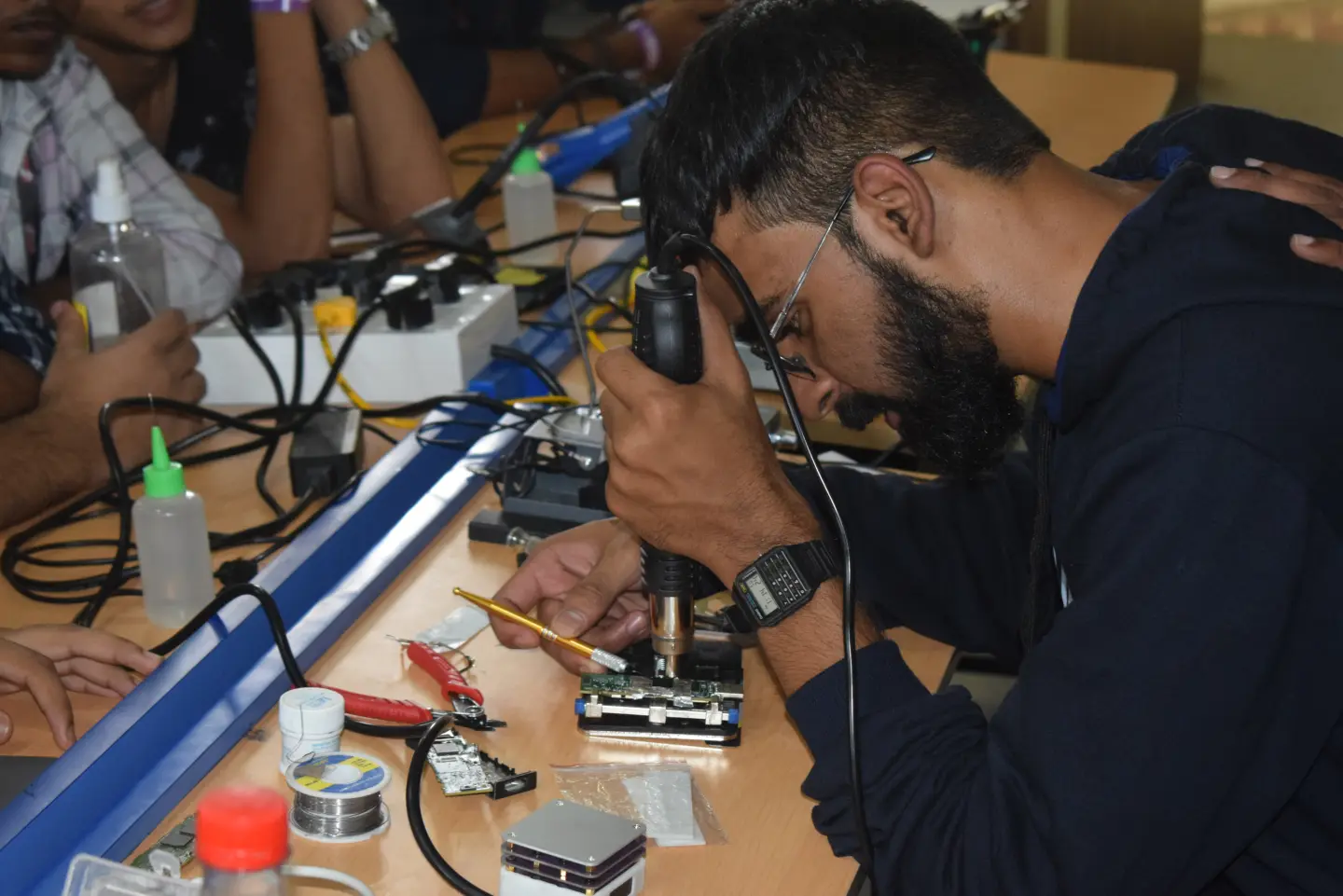 Someone bent over a workbench with a hot-air rework tool, working on a small circuit board in a jig while others lean in to watch