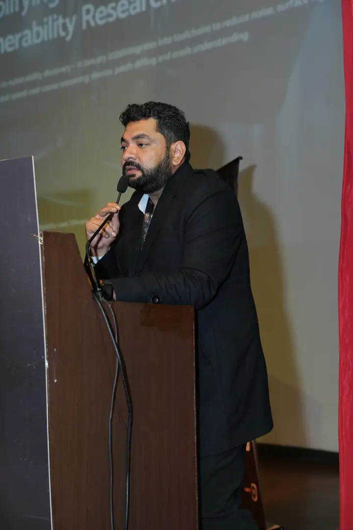 A speaker behind a wooden lectern, one hand raised, title slide on the wall behind