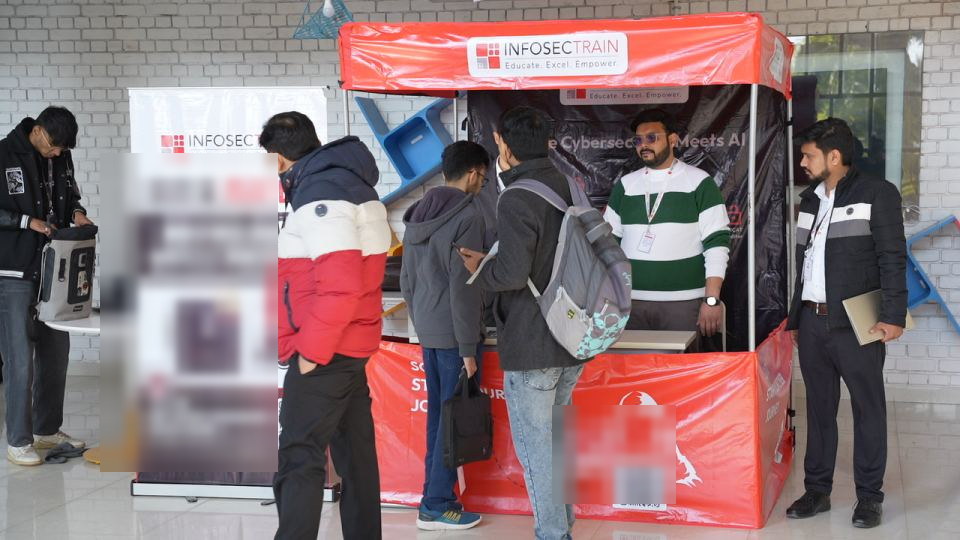 A sponsor's red pop-up stand at an indoor event, several attendees gathered at the counter talking with the two people staffing it, a printed roll-up banner standing to the left.
