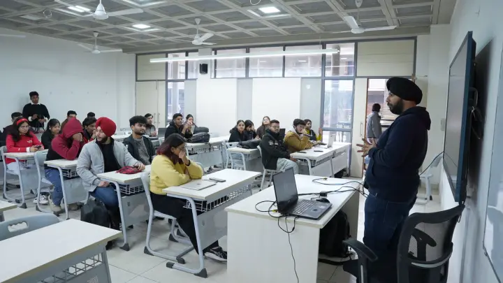 An instructor at the front of a bright classroom, a laptop open on the desk beside him, about twenty people at desks listening